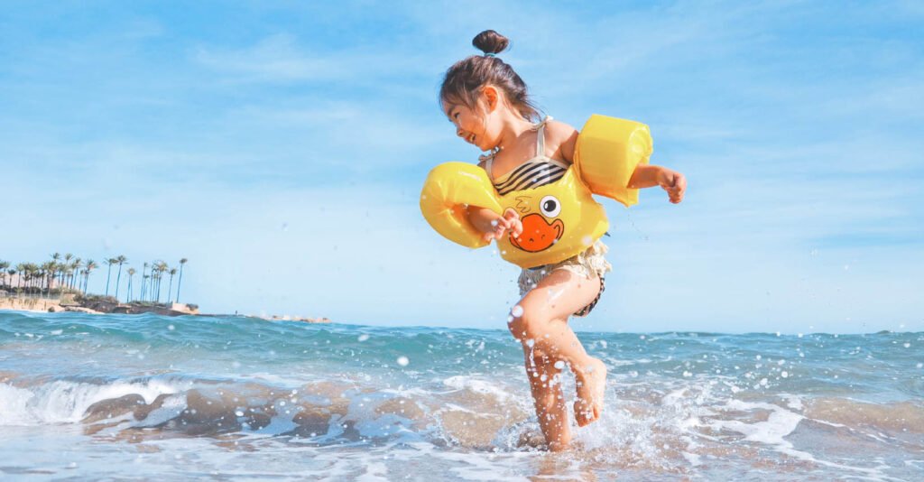 young child with yellow rubber ducky floaties playing in the water on a tropical beach with the best sun protection distant palm trees blue sky perfect day